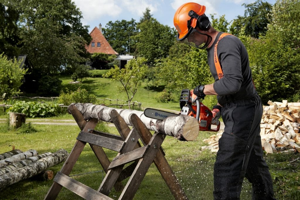 Landowner standing in his garden crosscutting a log lying on a sawhorse