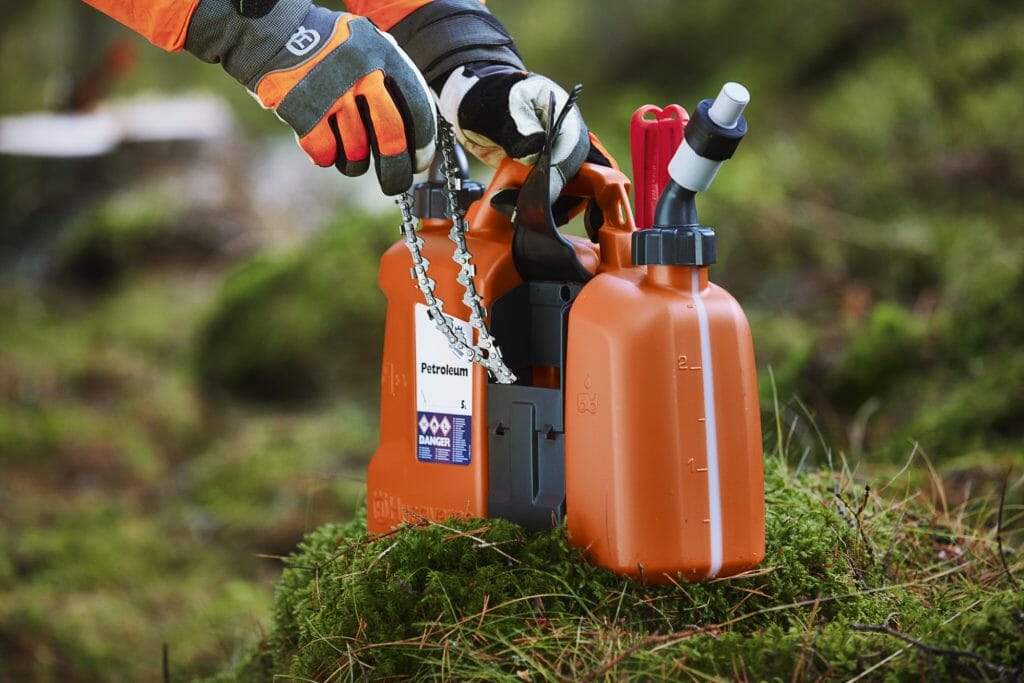 A spare chainsaw chain is pulled out of a compartment on a combi can for fuel and chain oil